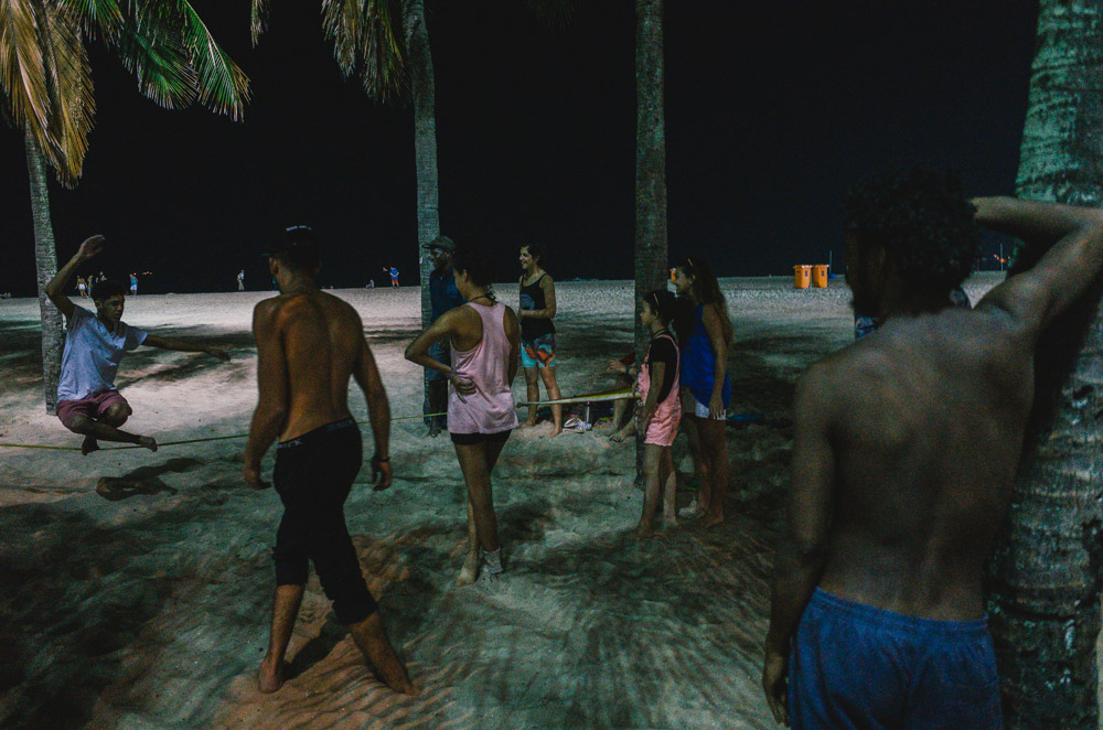 A guy slacklining on a line tied to trees on a beach at night as people watch.