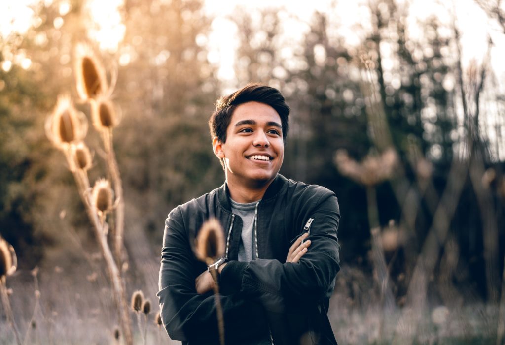 Portrait of a man with dark hair and spiked hair smiling that shows some plants in front of him out of focus and has a background out of focus