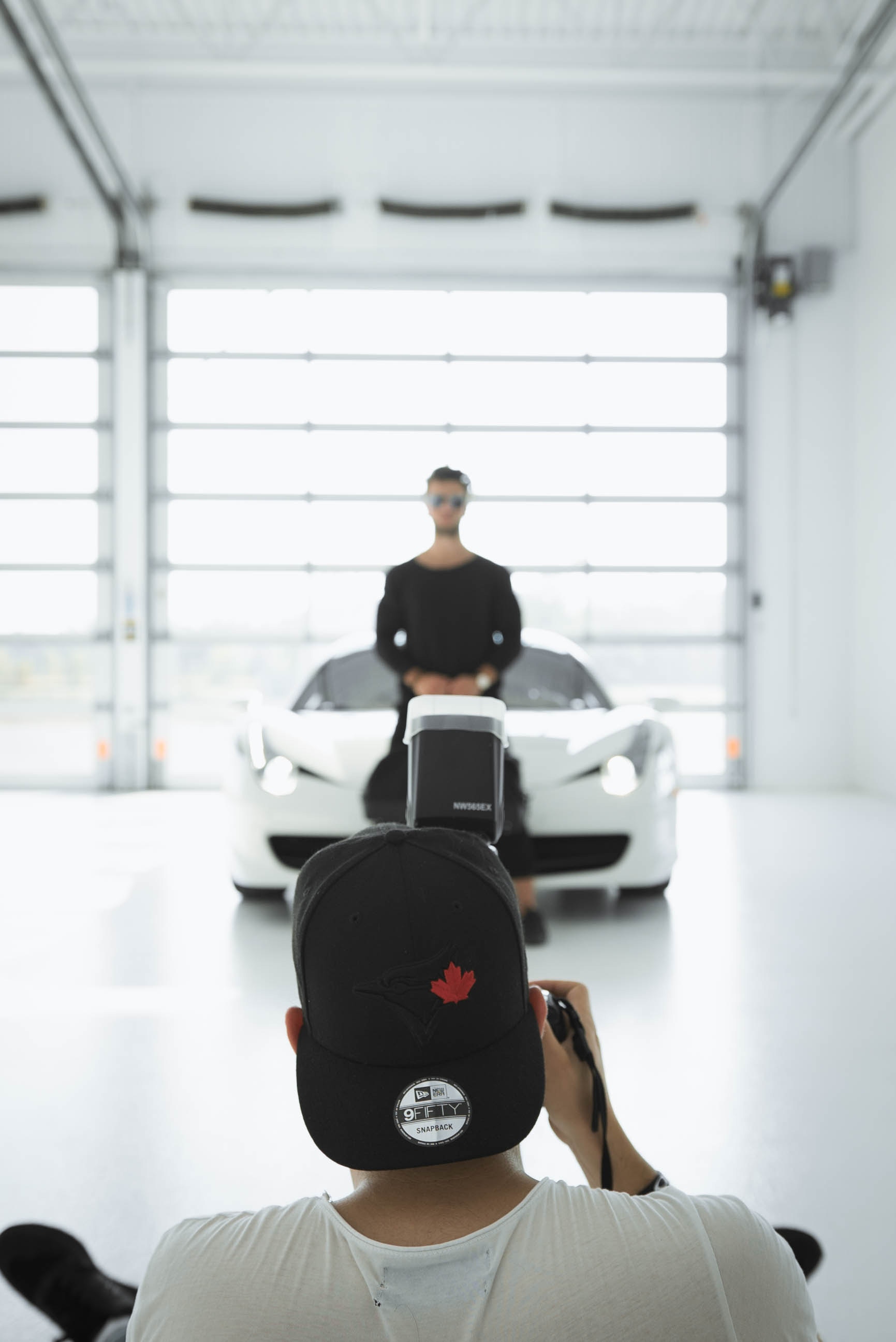 A photographer wearing a black backwards cap taking a portrait of a main standing in front of a white sports car.