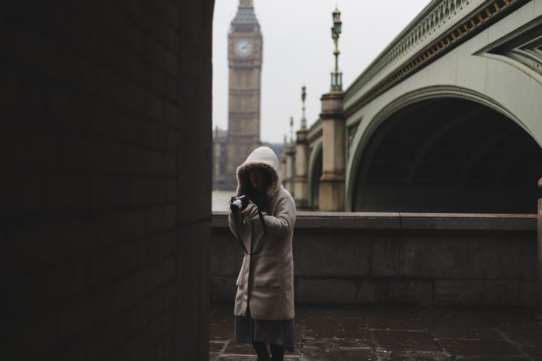A woman in a gray coat holding a camera with Big Ben and a bridge in the background.