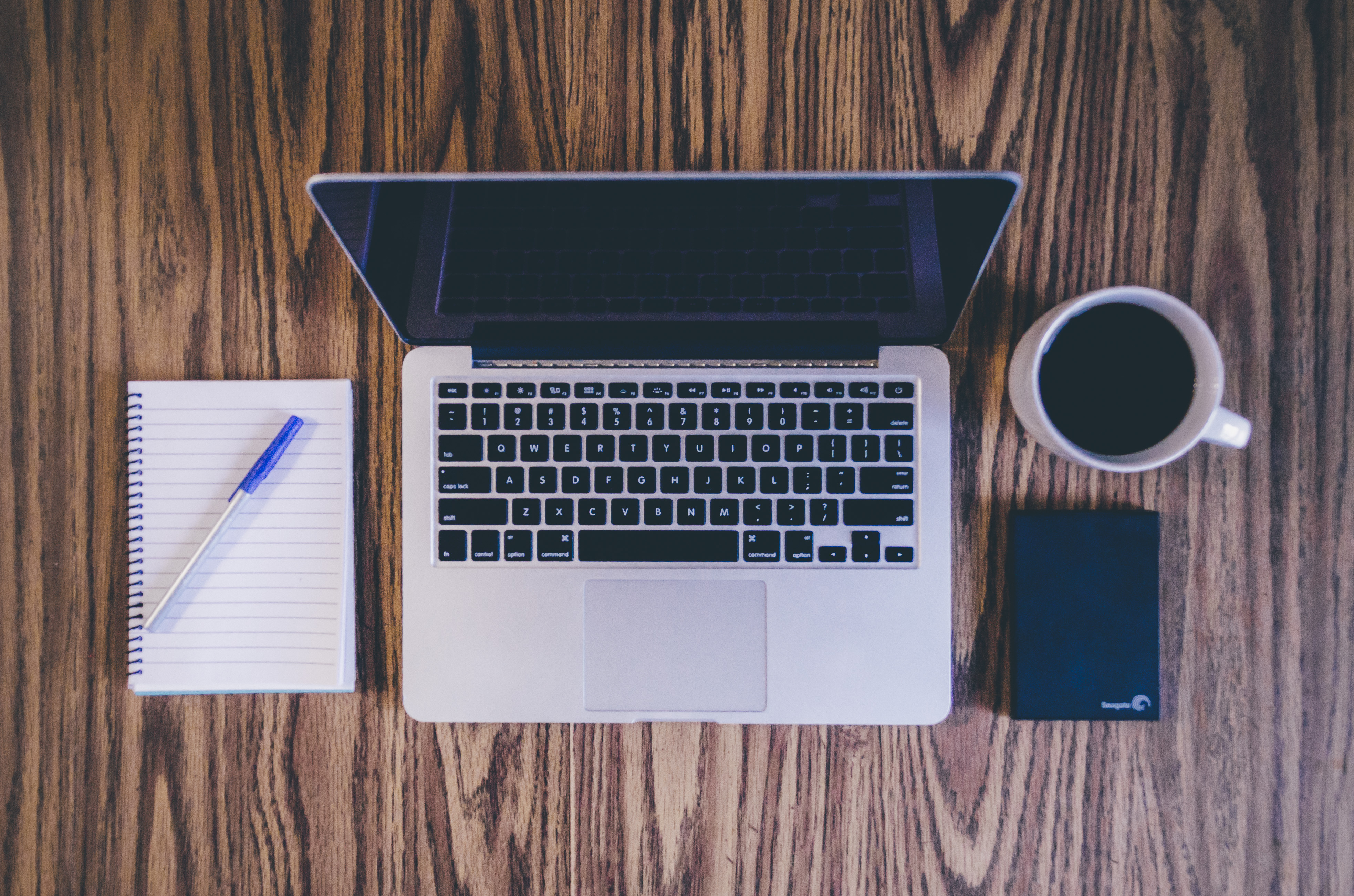 An Apple laptop computer, a notebook and pen, a hard drive, and a cup of coffee on a wood desk.