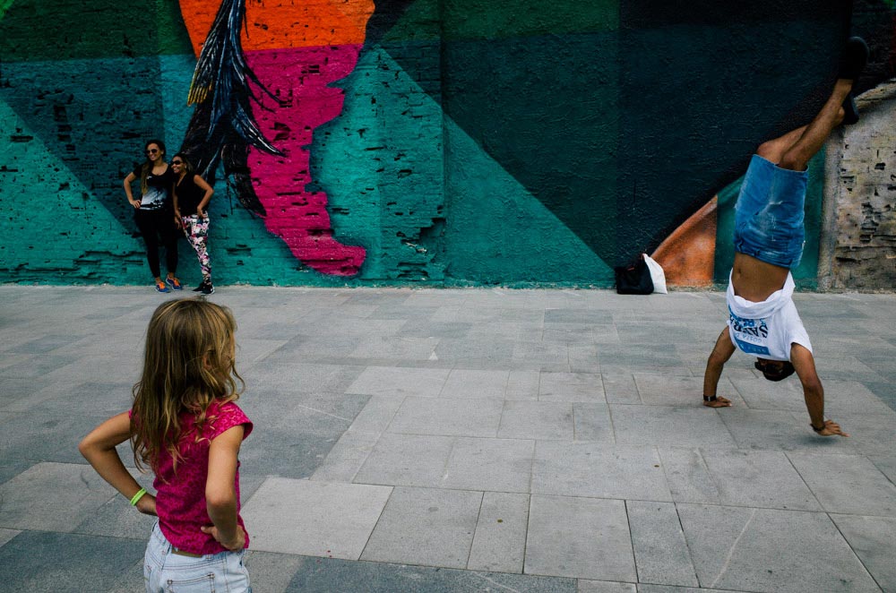 People in front of a map mural; two women posing for a photo, a young girl looking at a man doing a hand stand.