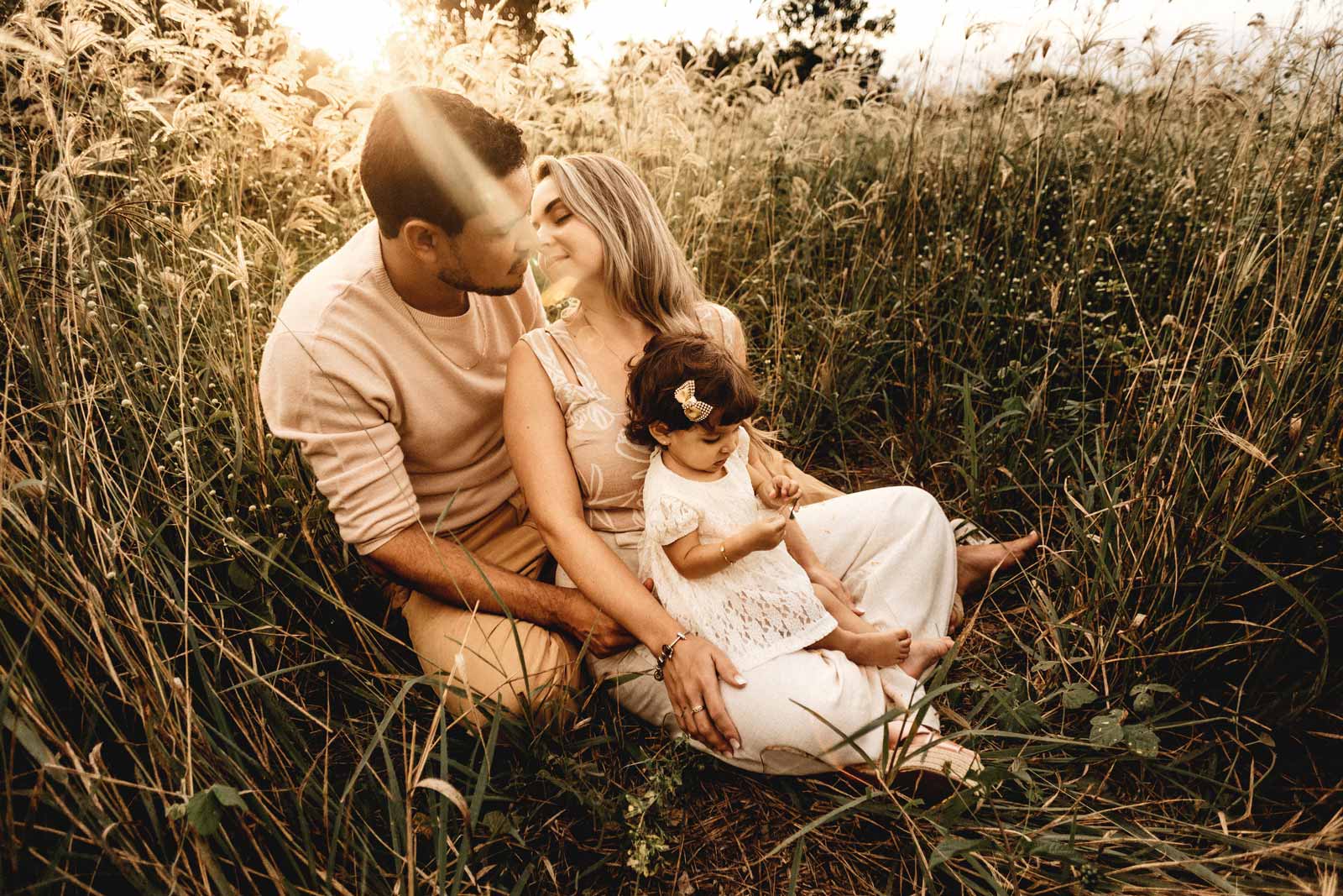 A man and woman kissing with a child in the woman's lap in a field with tall grass.