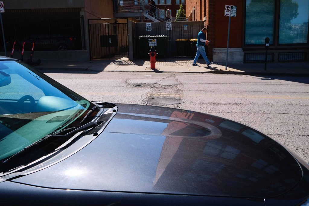 Man walking across street past an orange fire hydrant. In the foreground, there's a dark gray car.