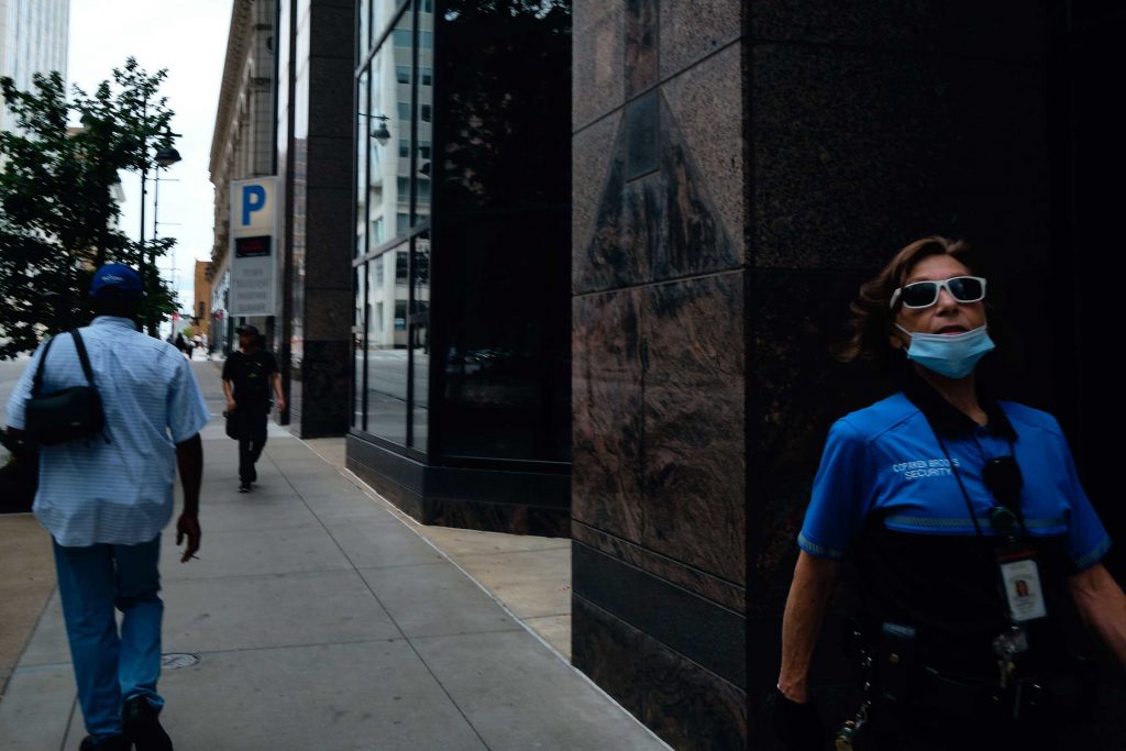 A woman security guard with white sunglasses on walking down a sidewalk with other pedestrians behind her.