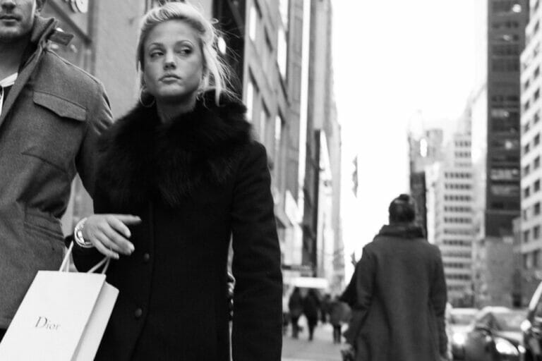 Blonde woman walking down sidewalk carrying Dior bag hand in hand with a man.