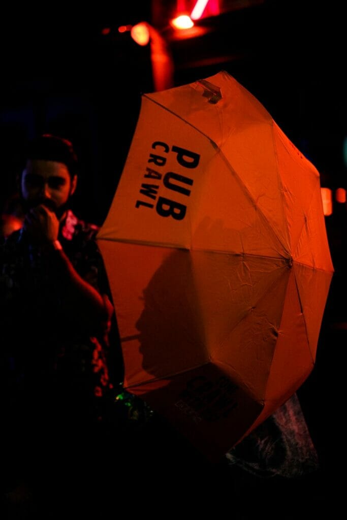 A man holding an umbrella at night that says 'PUB CRAWL' and a person's shadow on it.