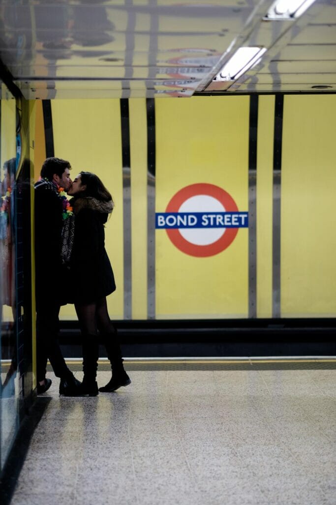 A young couple kissing in a subway station in front of a sign that says 'BOND STREET'.