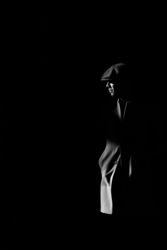 A man in a long coat and a newsboy cap in shadows.