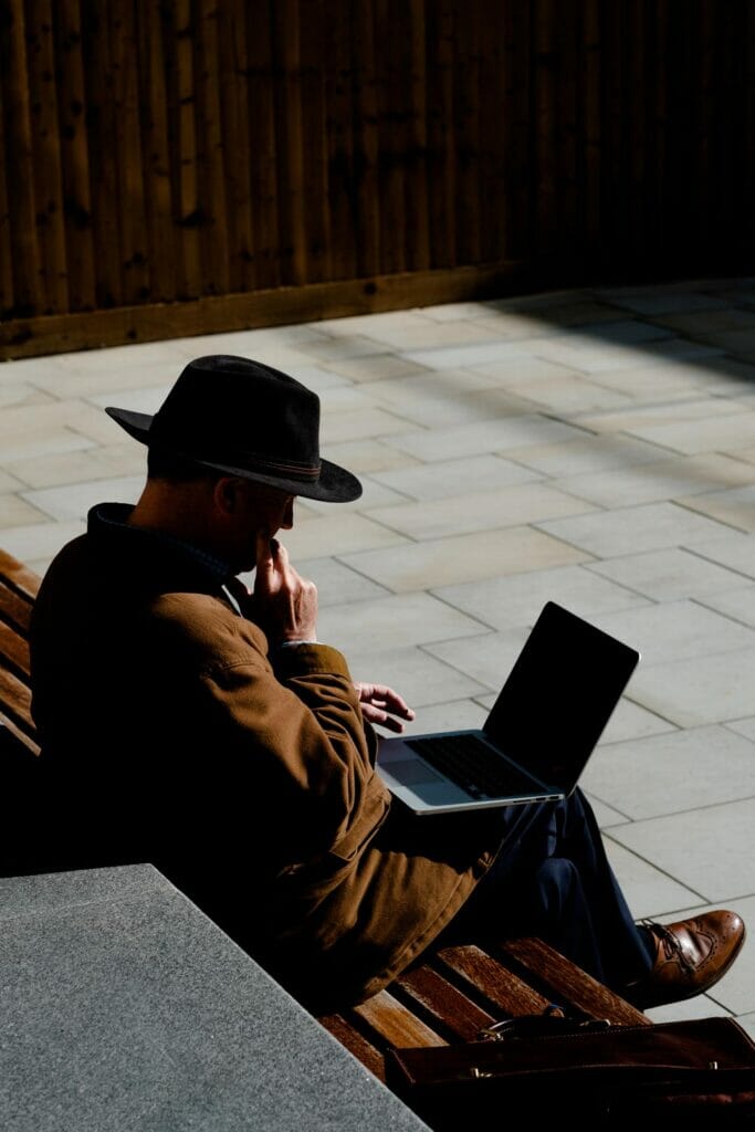 A man wearing a hat outside while looking at a laptop computer.