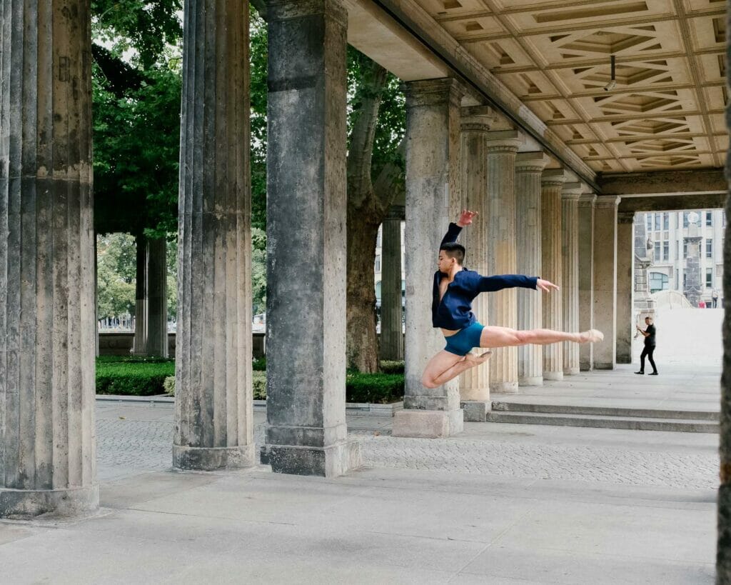 A ballerina man jumping under an old structure.