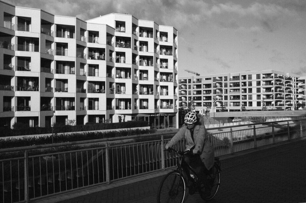 A person in a gray coat riding a bike on a riverfront with residential buildings.