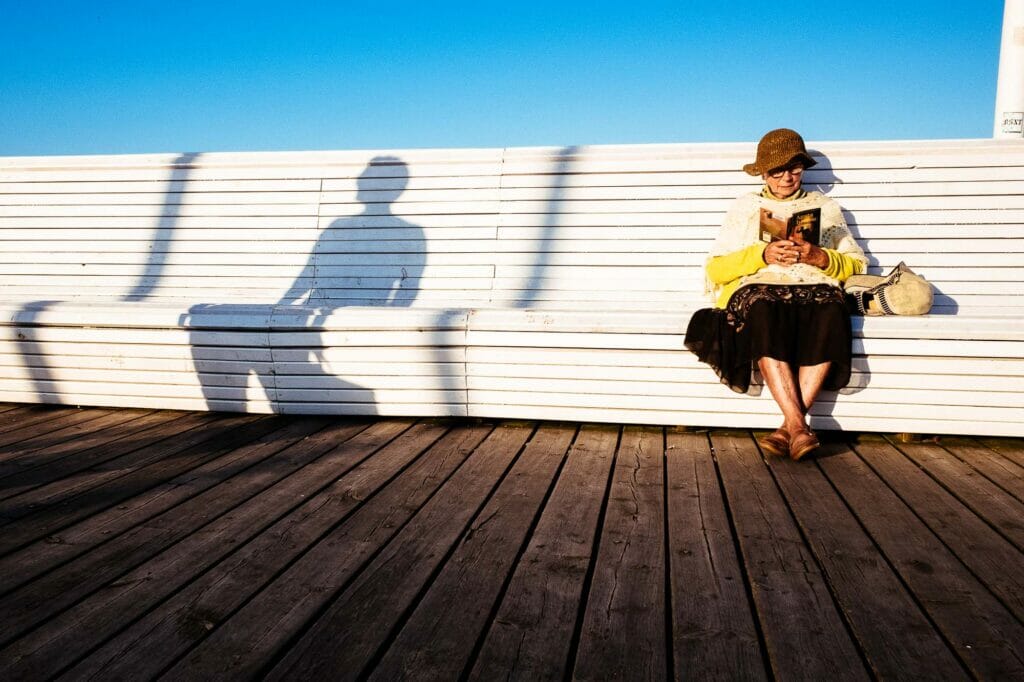 An old woman wearing a brown hat reading a book on a tall white bench.