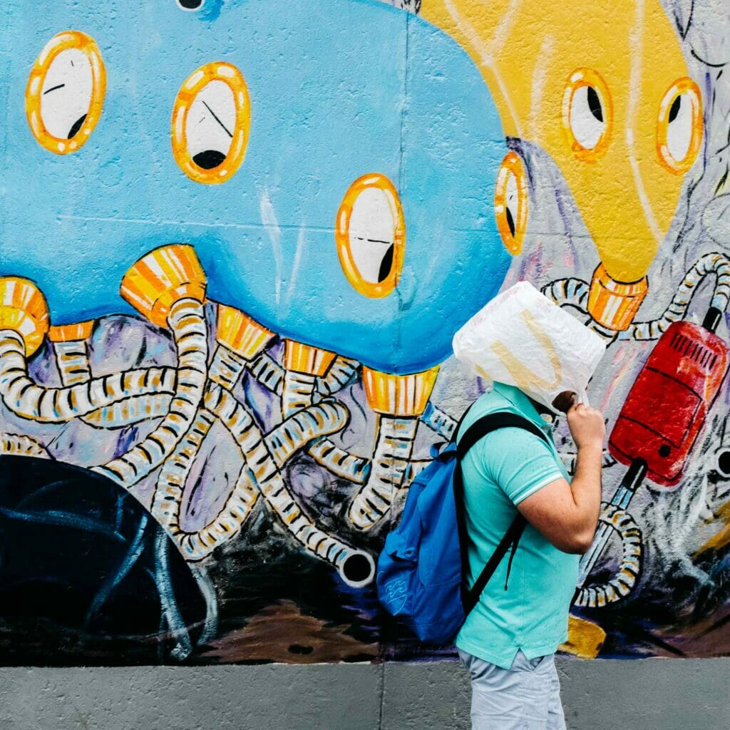 A man in a turquoise shirt and blue backpack holding a McDonald's bag in front of his face in front of a colorful mural.