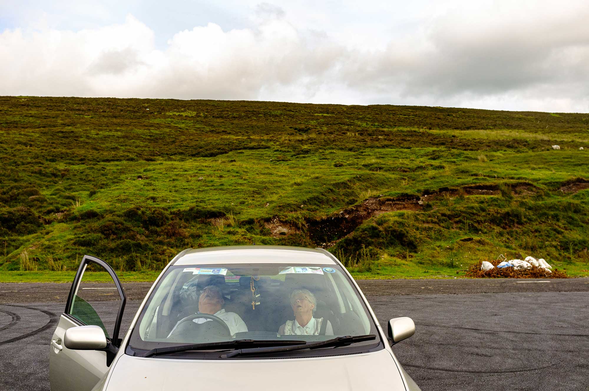 Old couple parked and asleep in a silver car in front of a lush green hill on a cloudy day.