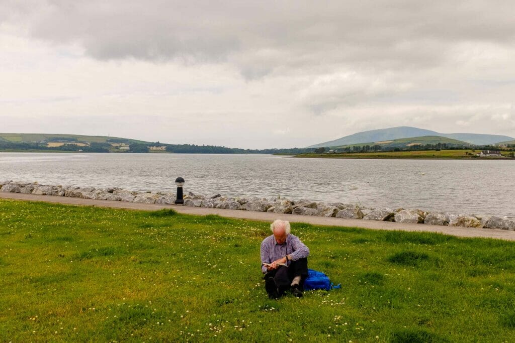 Older man with white hair reading a book by a lake on an overcast day