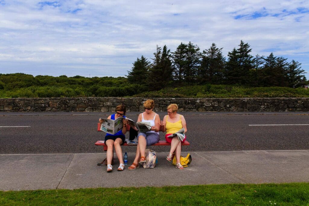 Three women on a bench in front of a street reading newspapers