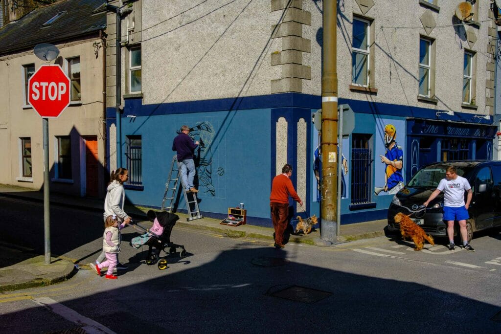 Street corner with two people walking dogs, a woman and young girl pushing a stroller, and a man painting a wall