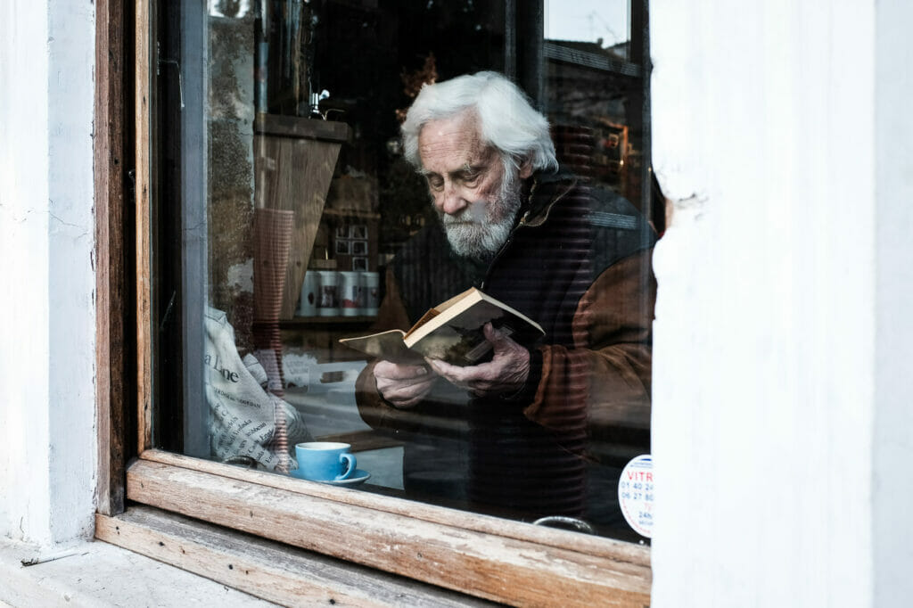 An old man reading a book through a shop window.