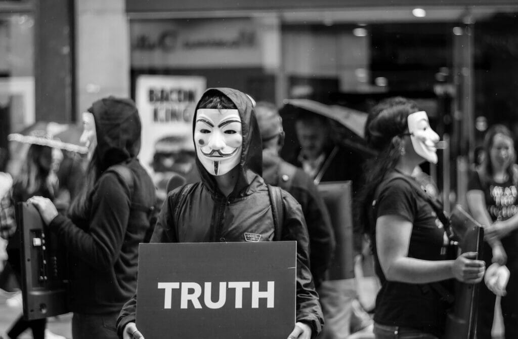 A protestor wearing a Guy Fawkes mask holding a sign that says 'TRUTH'.