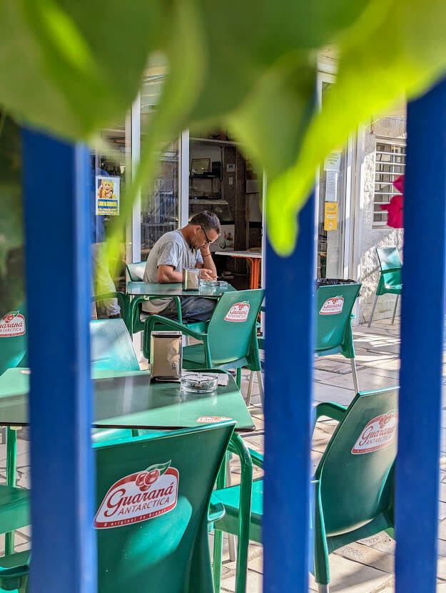 A man framed sitting in an outdoor cafe with green chairs and tables.