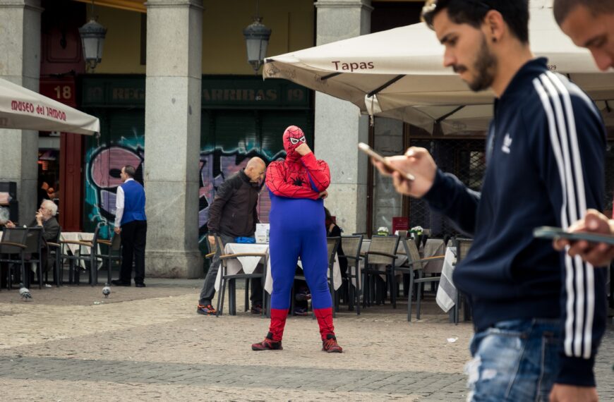 A person dressed as spider man in a public place with a man holding his phone walking in front.