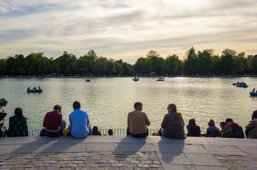 People sitting on steps in front of a pond with people rowing boats.