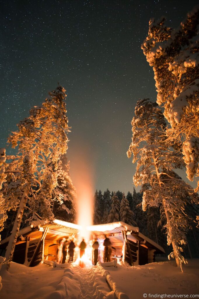 A group of people around a fire in a snowy place with tall fir trees.