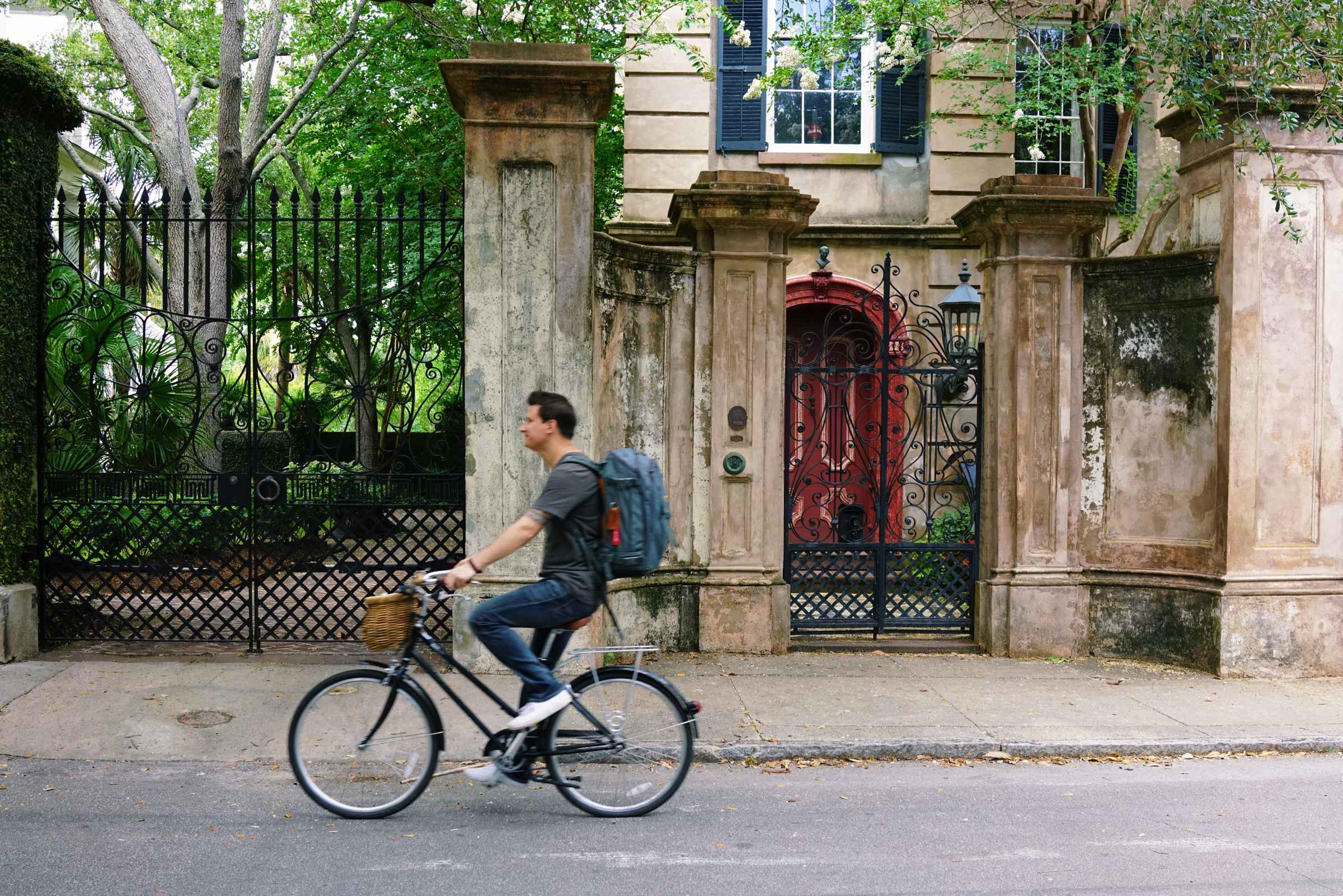 A man wearing a backpack riding on a bike in front of an old building with a gate.