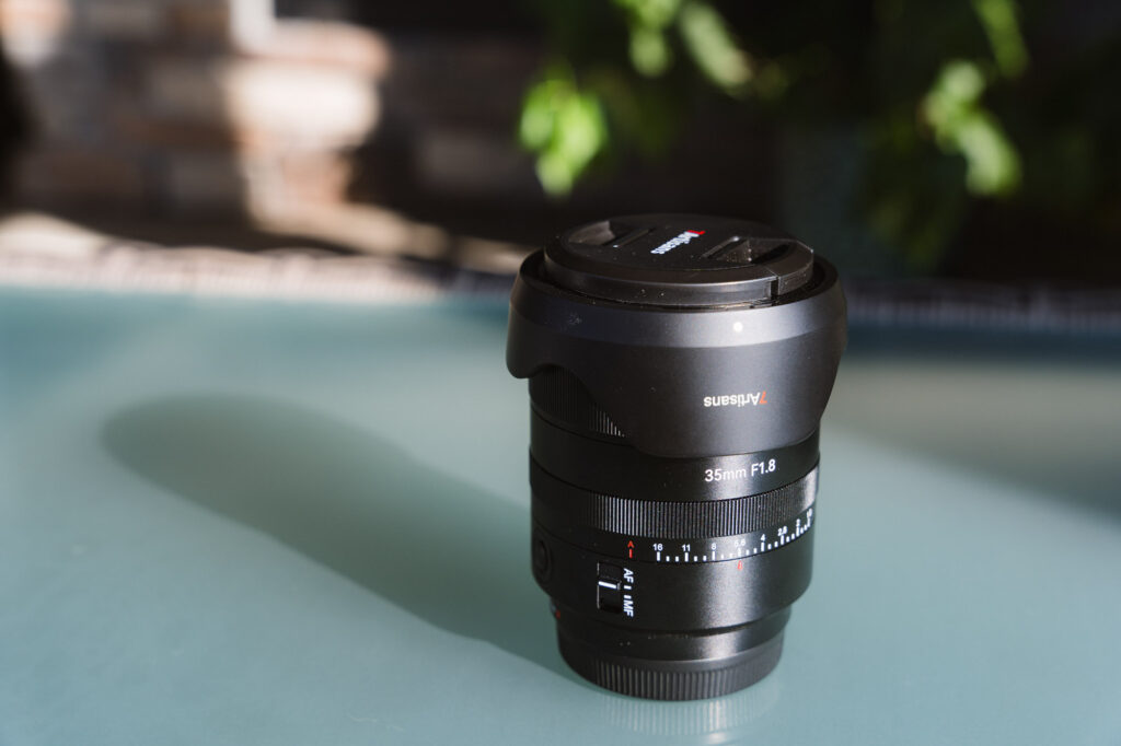 The 7Artisans AF 35mm f/1.8 Full Frame Lens on a glass table with its lens cap on and lens hood on backwards.