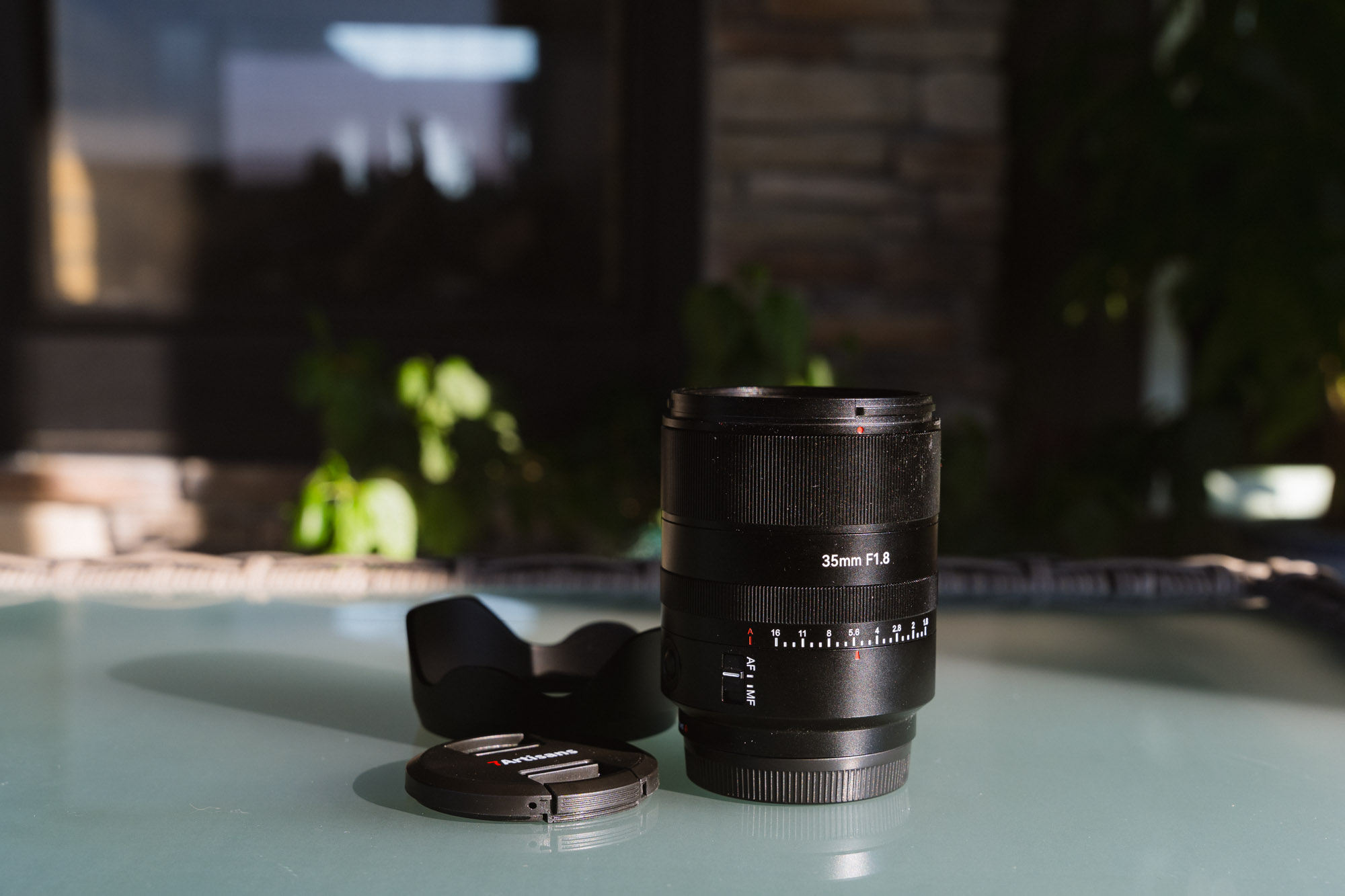 The 7Artisans AF 35mm f/1.8 Full Frame Lens on a glass table next to its lens hood and lens cap.