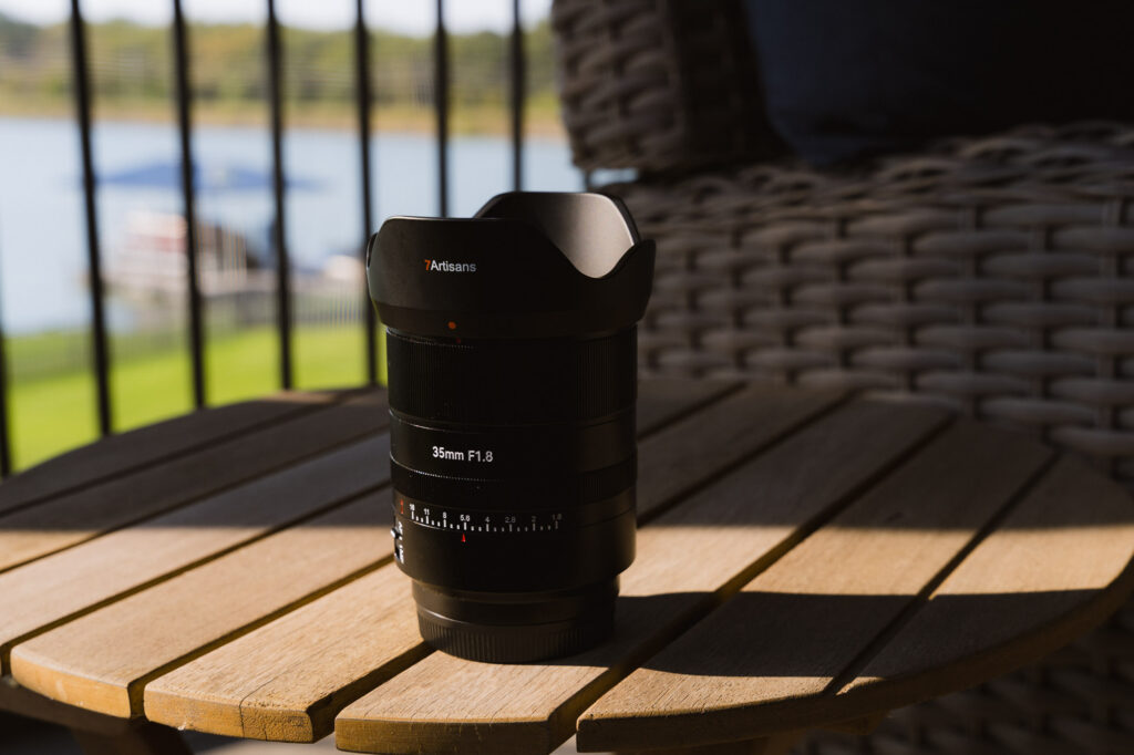 The 7Artisans AF 35mm f/1.8 Full Frame Lens with its lens hood on on a wood table in the sun.